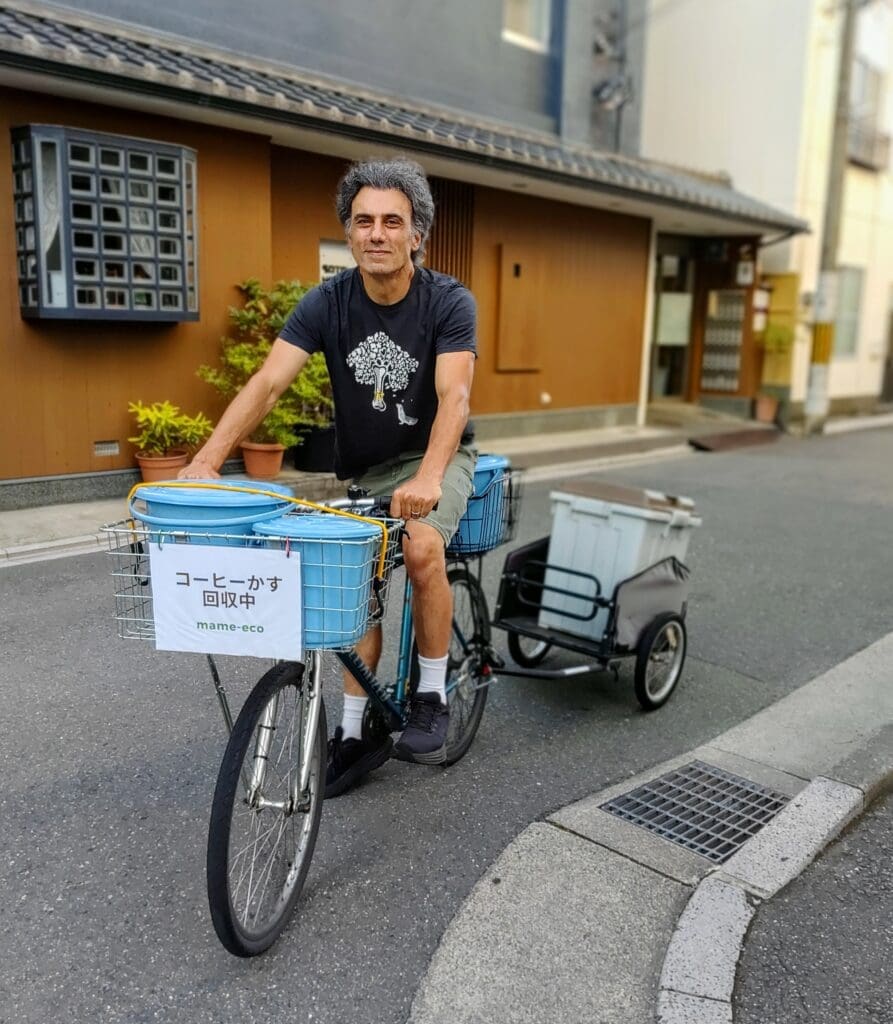 Gary from the mame-eco project riding a mountain bike with a trailer through a quiet Nishijin street in Kyoto, with blue coffee grounds collection buckets in the front and rear baskets.