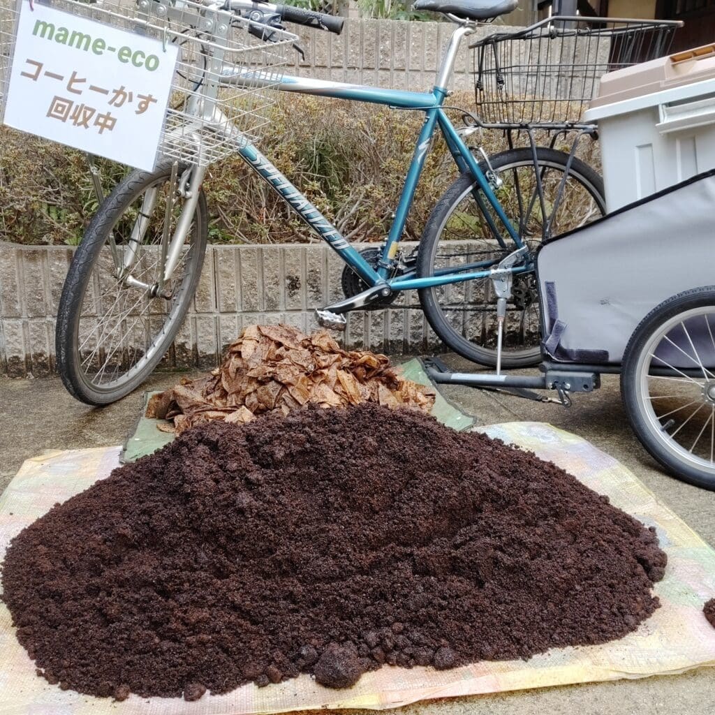 A mountain bike with a "mame-eco: Coffee Grounds Collection in Progress" sign, parked behind piles of used coffee filters and a large heap of coffee grounds in the foreground.
