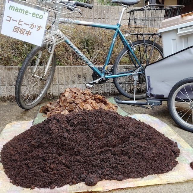 「mame-eco コーヒーかす回収中」の看板がついたマウンテンバイクと、手前に積み上げられた使用済みのコーヒーフィルターとコーヒーかすの山。