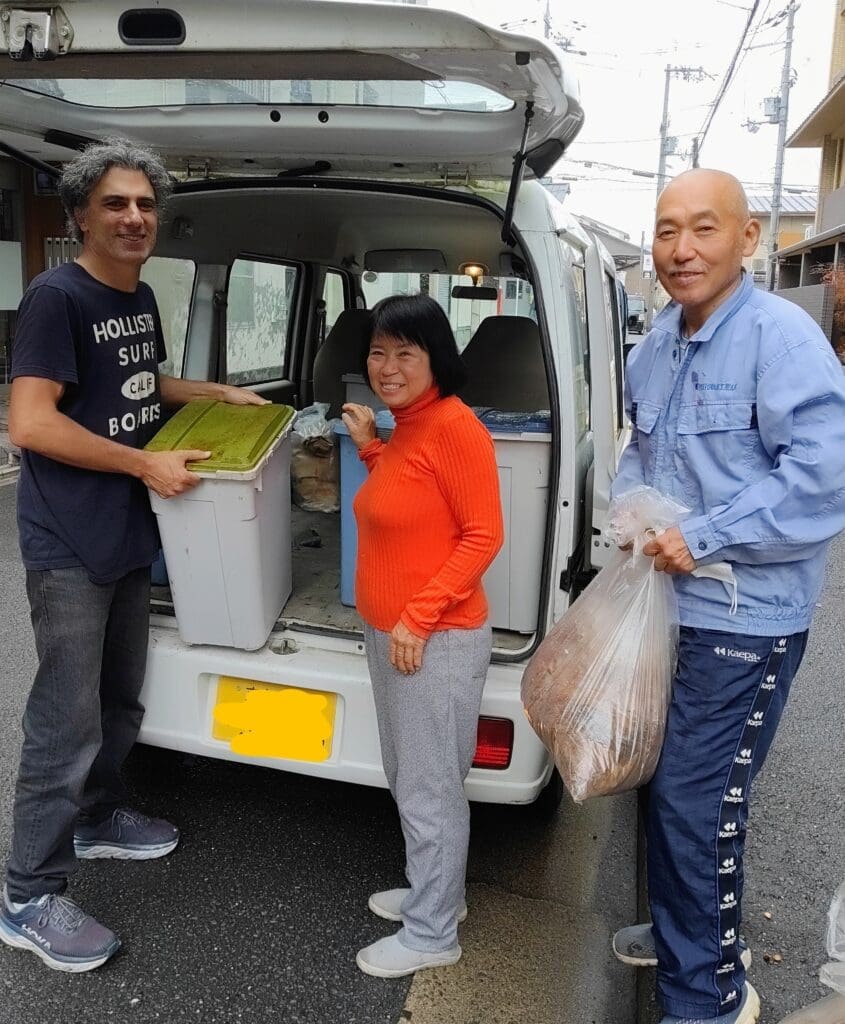 Gary from the mame-eco project loading a large bucket of coffee grounds into a van while a Kameoka farm owner and a fellow farmer look on.