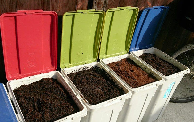 Four large buckets filled with collected coffee grounds next to an old wooden Kyoto house, with sunlight streaming over the grounds and colorful open lids.