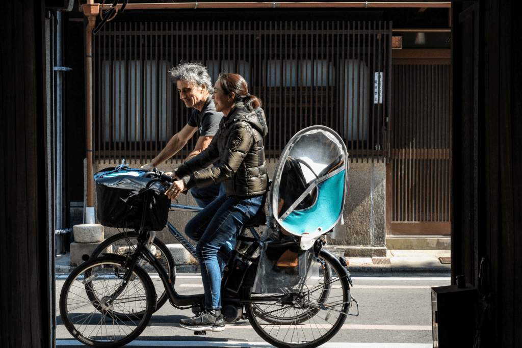 京都の静かな通りで、京町家を背景に並ぶmame-eco(マメエコ)のGaryとJunko。二人の自転車にはコーヒーかす回収用の青いバケツが載っています。